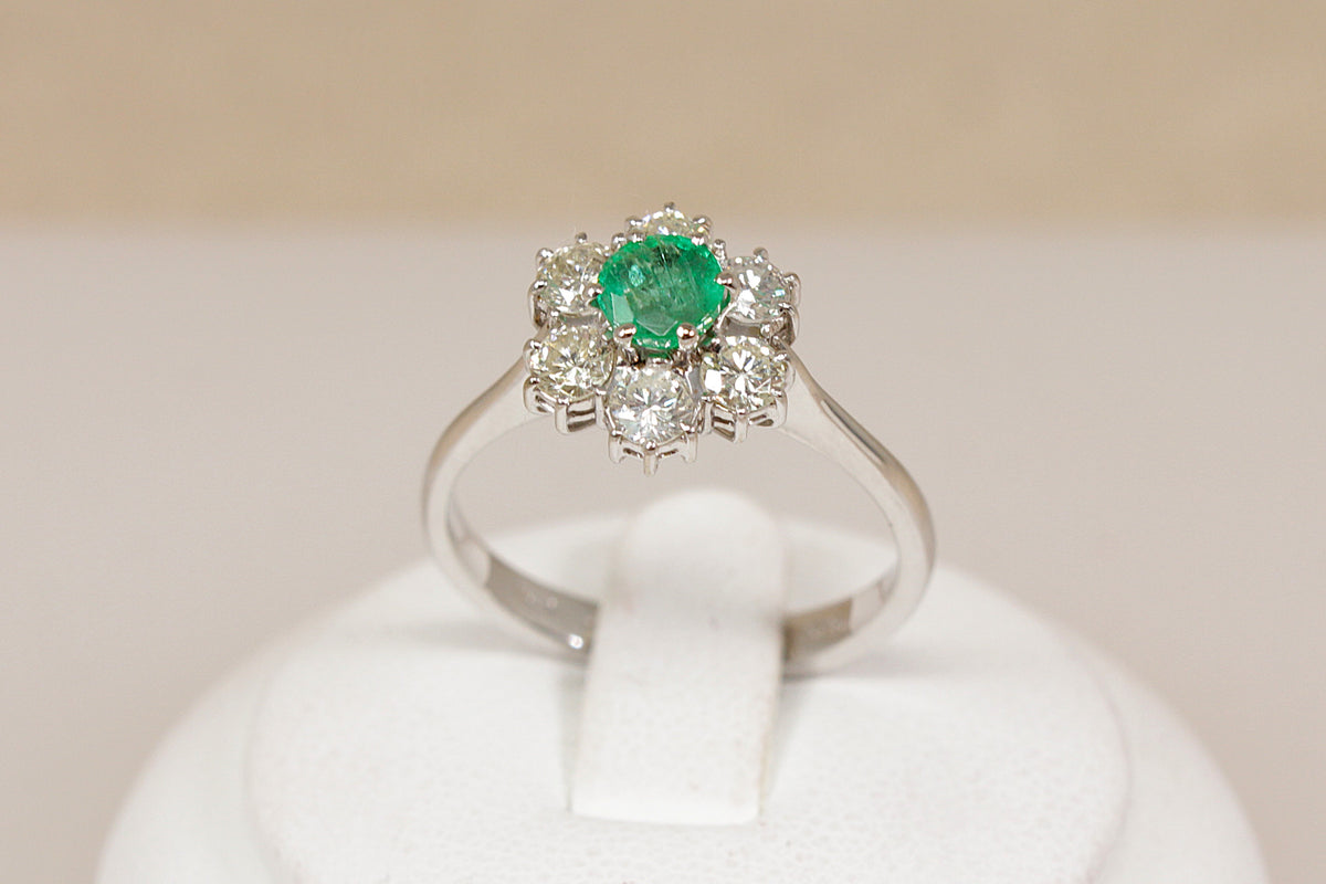 A 18ct white gold cluster ring featuring a round green emerald center stone surrounded by small diamond gemstones, displayed on a white ring stand against a soft beige background.