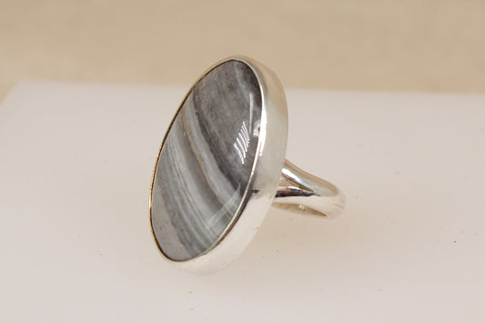 A silver ring featuring a large, oval-shaped cabochon banded agate gemstone with distinct gray and white banding, set against a neutral beige and white background.
