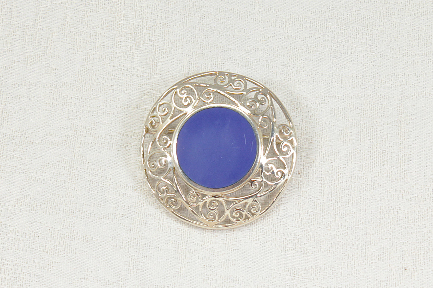 A circular silver filigree brooch featuring a smooth, vibrant blue lapis lazuli round  center gemstone, displayed on a textured off-white fabric background.