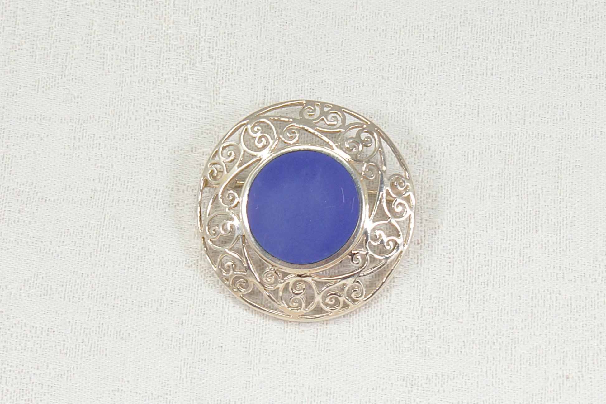 A circular silver filigree brooch featuring a smooth, vibrant blue lapis lazuli round  center gemstone, displayed on a textured off-white fabric background.