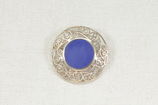 A circular silver filigree brooch featuring a smooth, vibrant blue lapis lazuli round  center gemstone, displayed on a textured off-white fabric background.