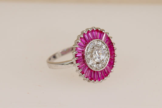 A close-up of a sterling silver ruby and white crystal ring with a central clear gemstone and a surrounding halo of pink ruby stones.