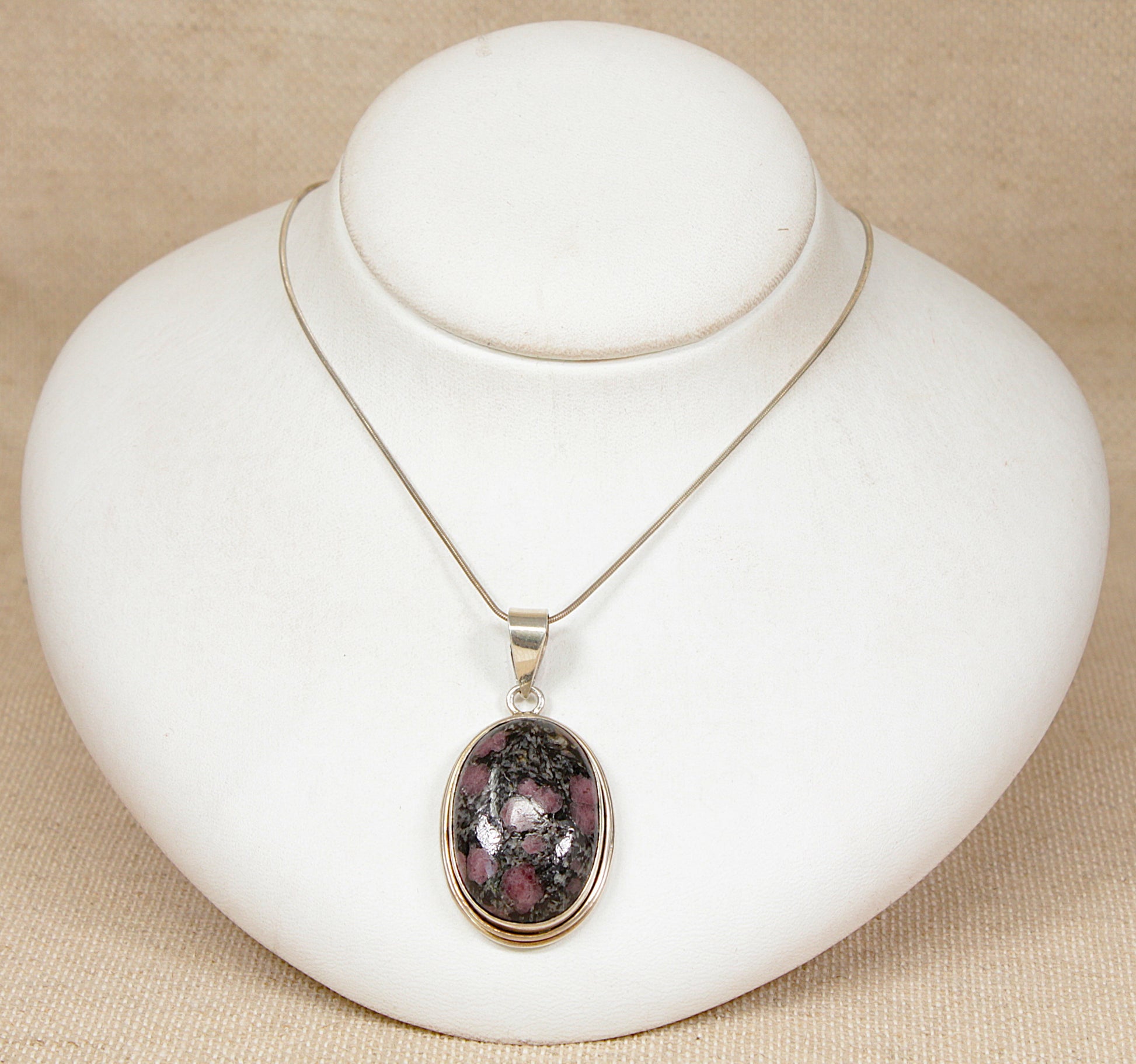 An oval pendant necklace with a spotted jasper agate gemstone silver bezel pendant necklace displayed on a white jewelry bust against a beige textured background.