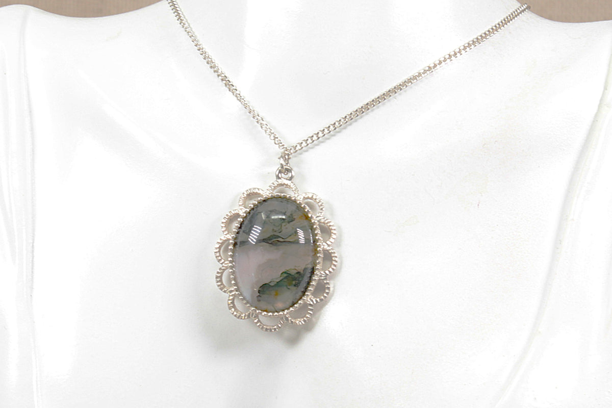 A silver chain necklace with an oval pendant featuring a moss agate  gemstone, set in a silver filigree frame, displayed on a white mannequin bust.