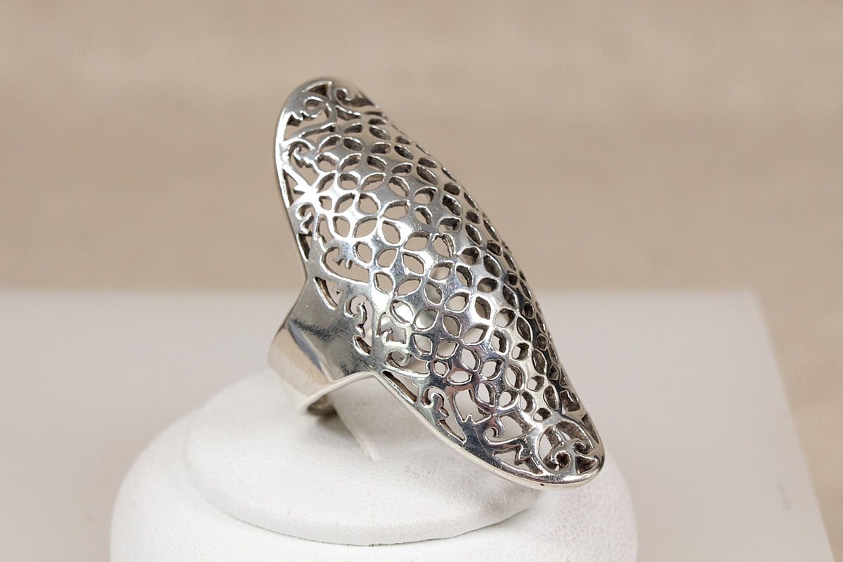 A wide, sculptural silver statement ring with an intricate openwork net-like pattern, displayed on a white circular stand against a soft beige background.