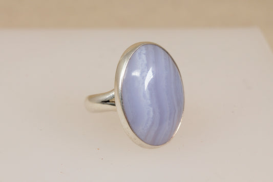 A silver statement ring featuring a large oval cabochon blue lace agate gemstone with banding, displayed on a neutral cream background.