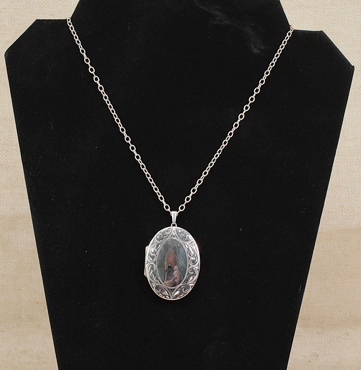 A silver oval locket necklace with engraved floral detailing, displayed on a black velvet jewelry bust against a beige fabric background.