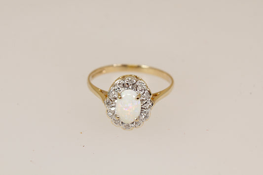 A yellow gold ring featuring a central oval white opal surrounded by a halo of small diamond gemstones, set against a plain light beige background.
