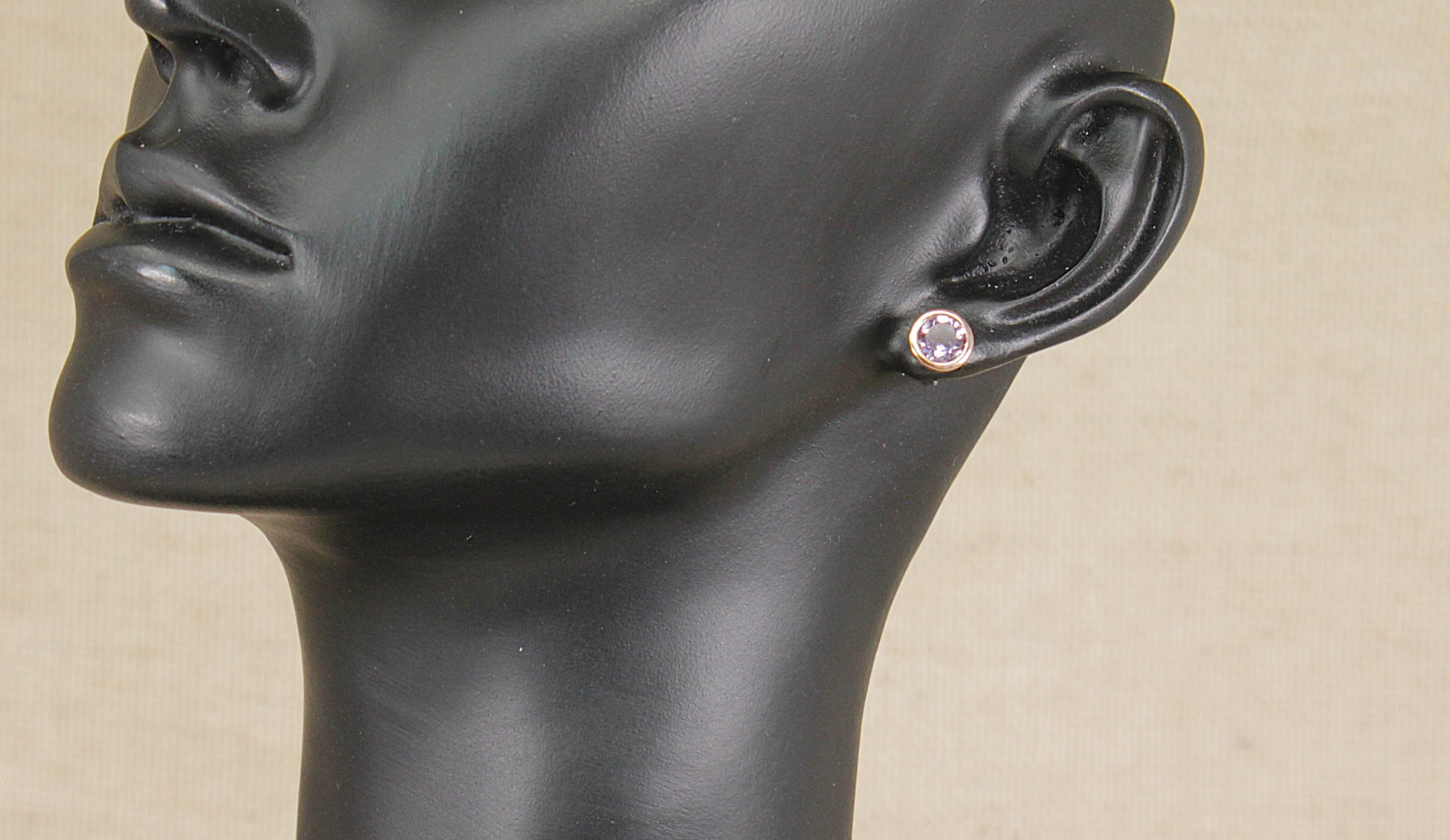 A close-up of a black mannequin head and neck wearing a small round Clogau silver amethyst and gold stud earring on a beige background.
