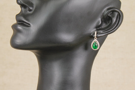 A single teardrop-shaped drop earring with a green center stone and a silver-toned halo, displayed on a black mannequin head against a light beige background.