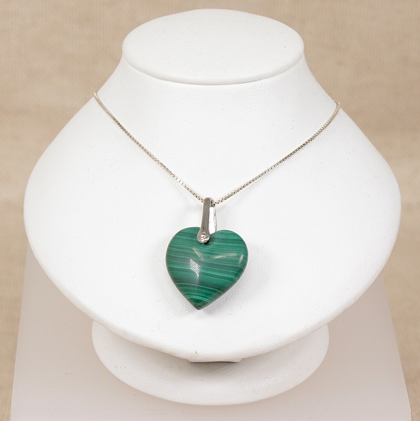 A heart-shaped green malachite pendant necklace on a silver chain, displayed on a white jewelry bust against a light beige background.