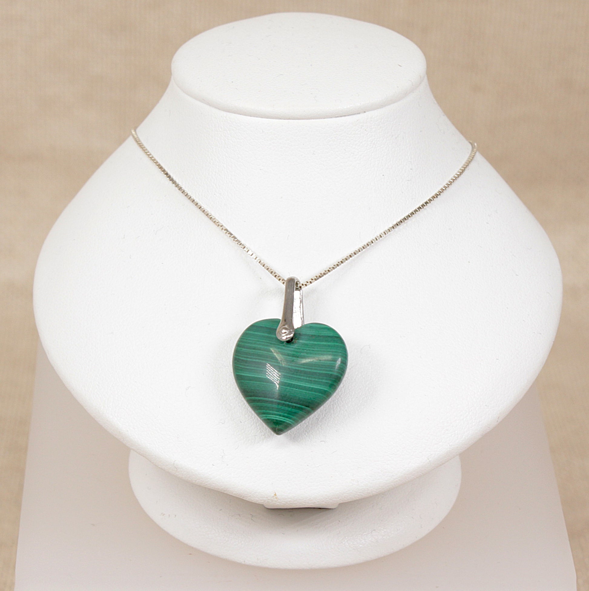 A heart-shaped green malachite pendant necklace on a silver chain, displayed on a white jewelry bust against a light beige background.