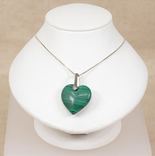 A heart-shaped green malachite pendant necklace on a silver chain, displayed on a white jewelry bust against a light beige background.