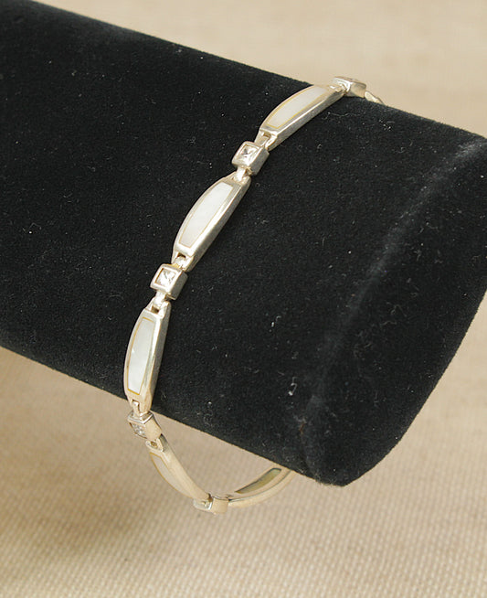 A sterling silver bracelet with alternating curved and square links, featuring mother of pearl white inlay spaced with cubic zirconia gemstones displayed on a black cylindrical jewelry stand against a beige fabric background.