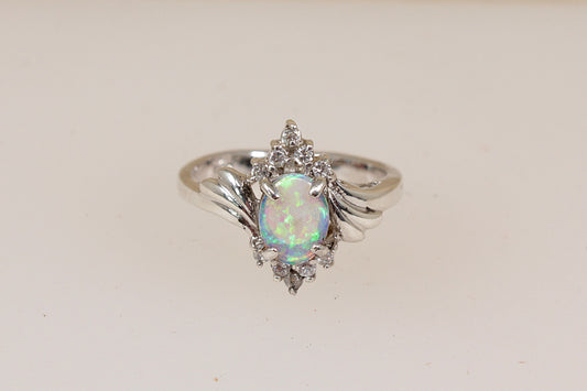 A sterling silver ring featuring an oval iridescent opal center stone surrounded by small clear accent gemstones and a wing-like halo, set against a plain light beige background.