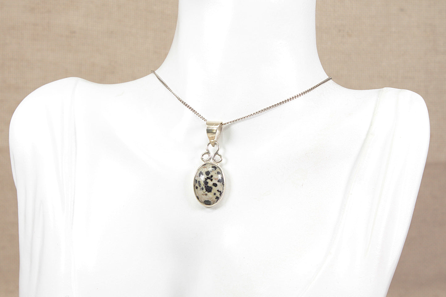 A silver chain necklace with an oval black and white leopard spotted jasper agate pendant, displayed on a white mannequin bust against a neutral beige background.