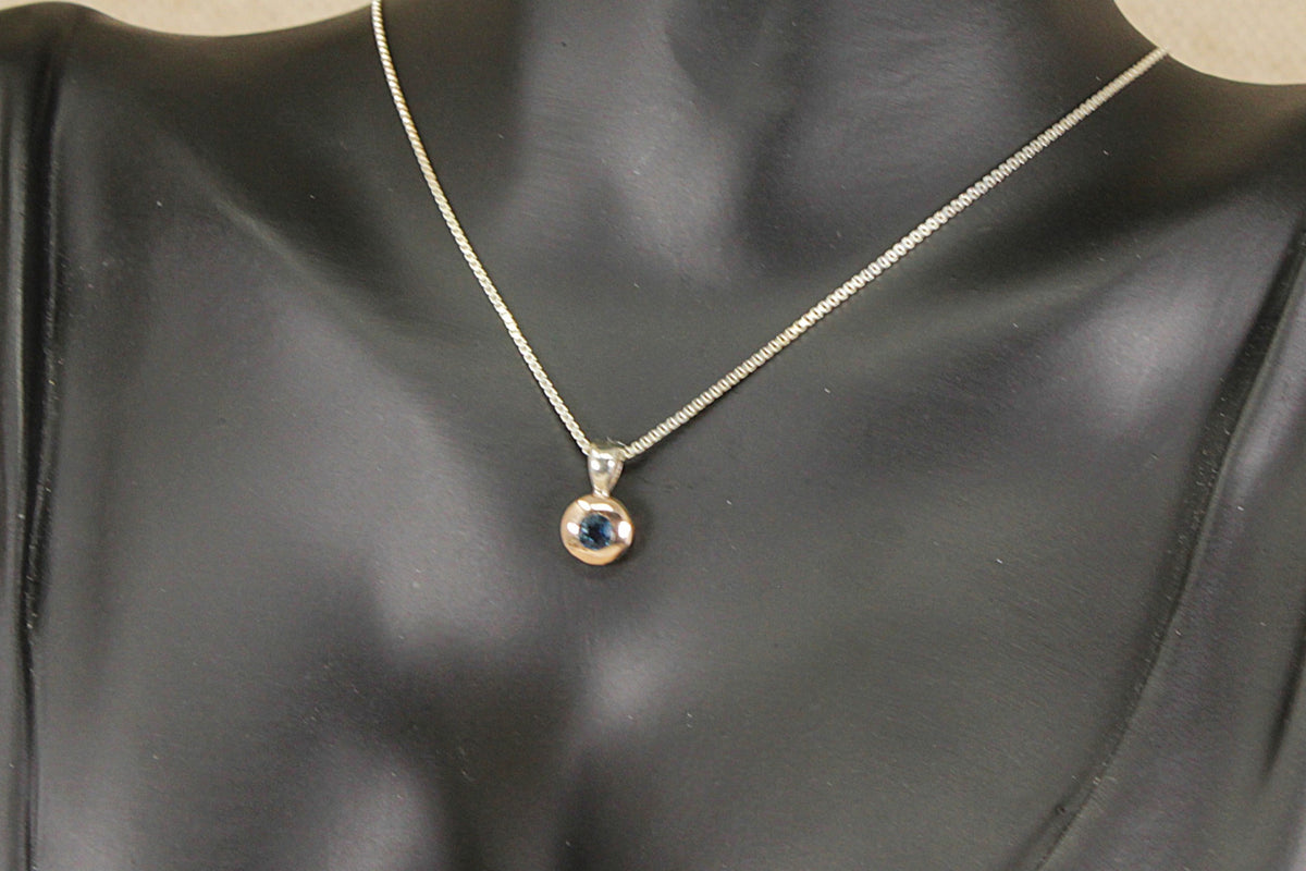 A delicate silver chain necklace with a small circular pendant featuring a dark blue sapphire gemstone, displayed on a black mannequin bust.