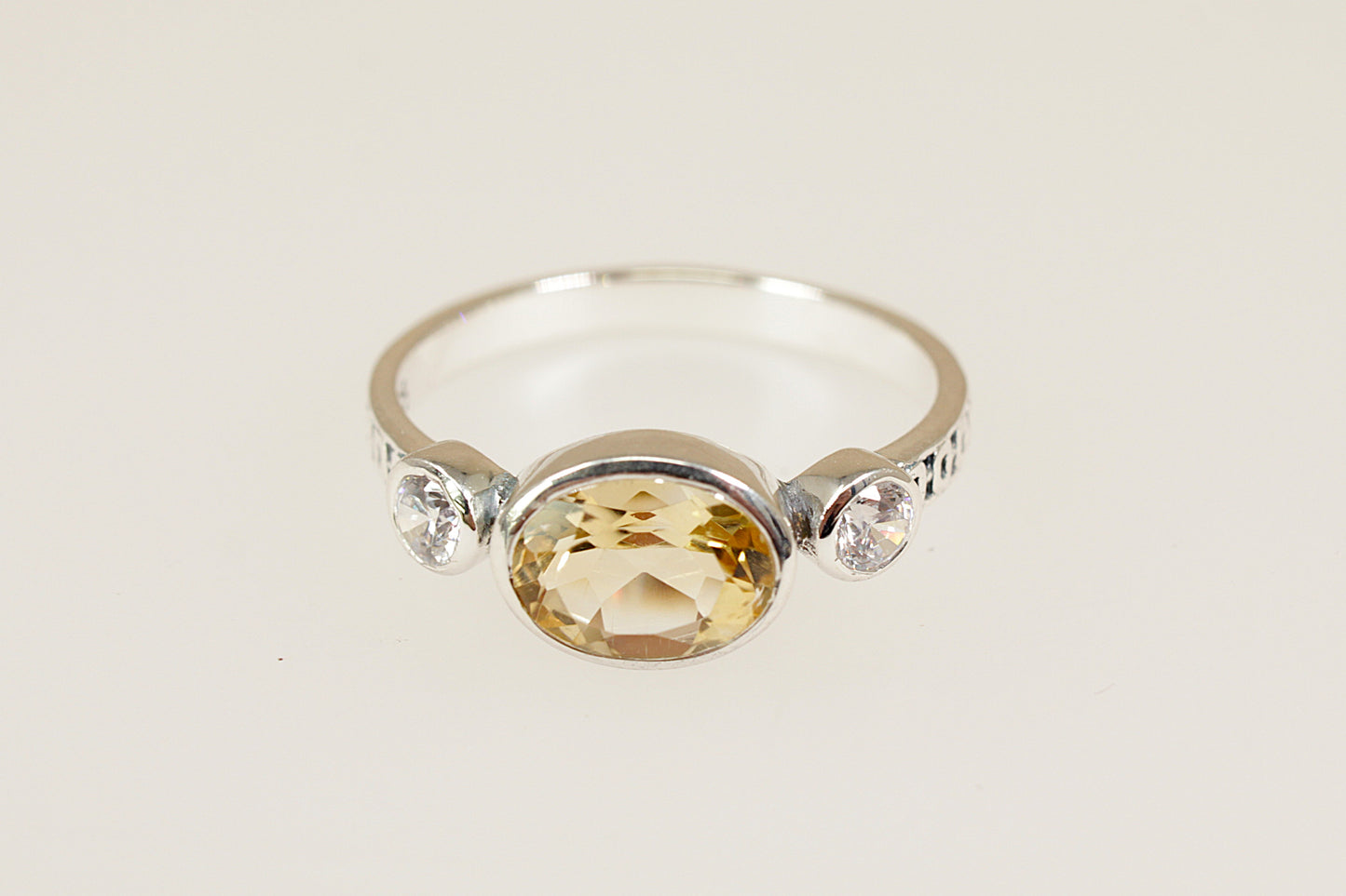 A silver ring featuring a large oval-cut yellow citrine center stone and two small clear accent stones on either side of the band, set against a plain light beige background.