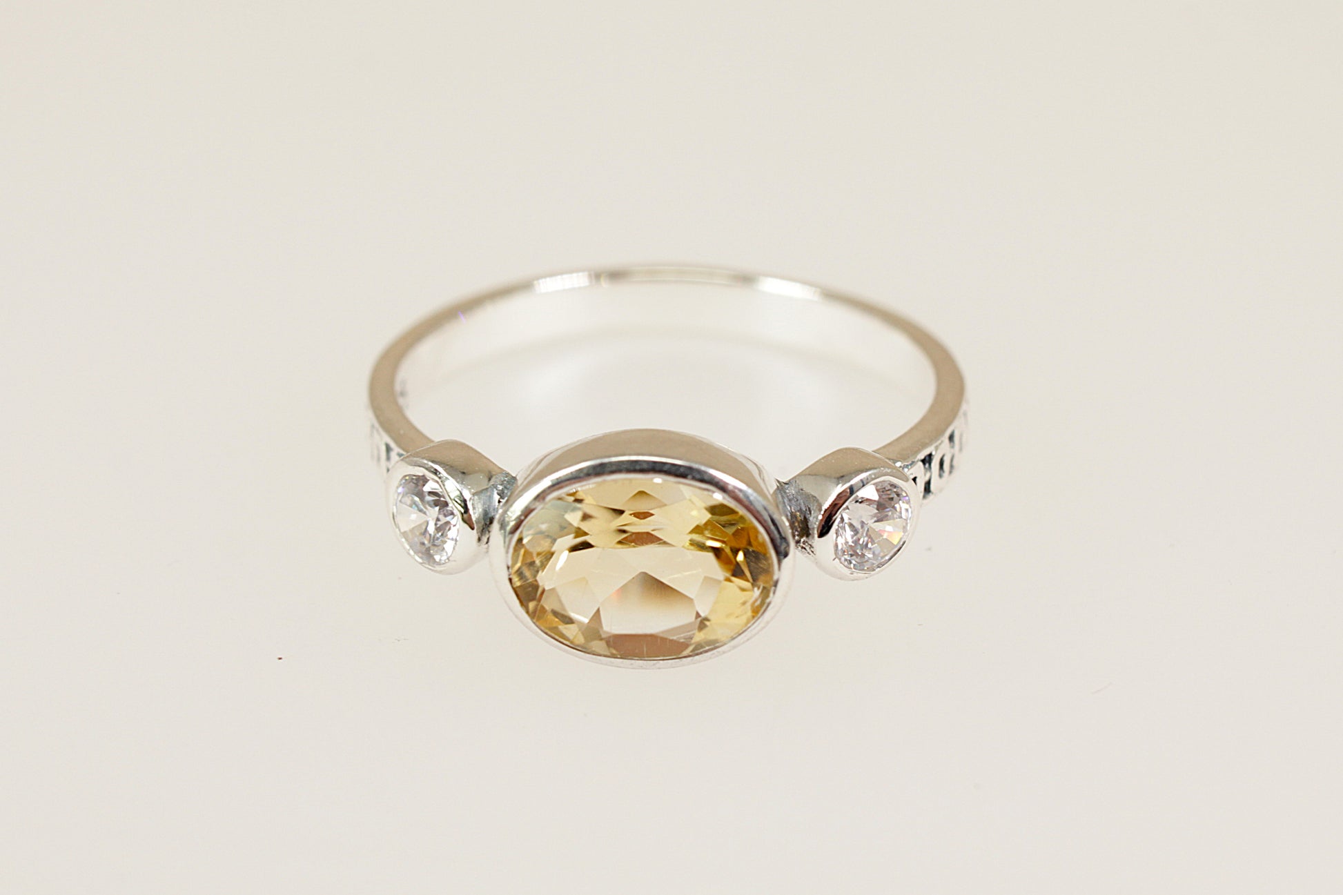 A silver ring featuring a large oval-cut yellow citrine center stone and two small clear accent stones on either side of the band, set against a plain light beige background.