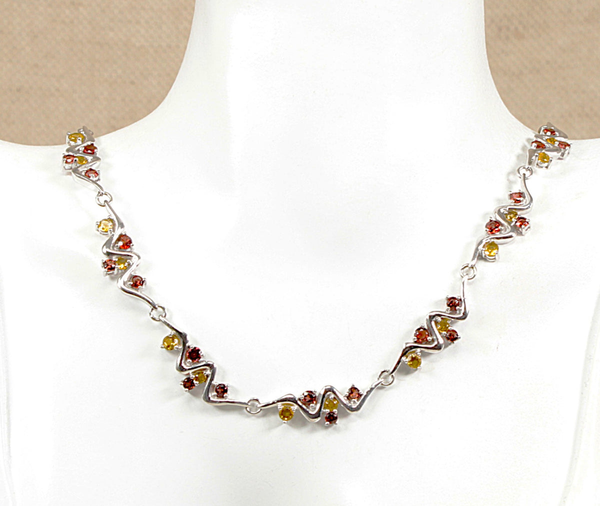 A silver chain necklace with small red garnet and yellow citrine gemstones, displayed on a white mannequin bust.
