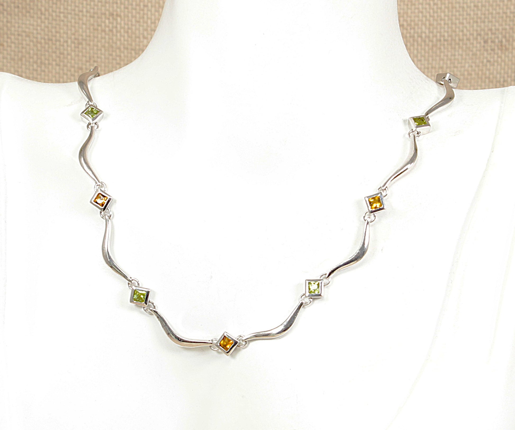 A silver wave necklace with alternating green peridot and citrine gemstone, displayed on a white mannequin bust.