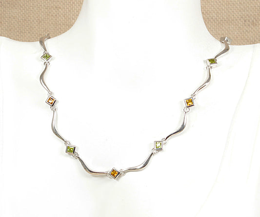 A silver wave necklace with alternating green peridot and citrine gemstone, displayed on a white mannequin bust.