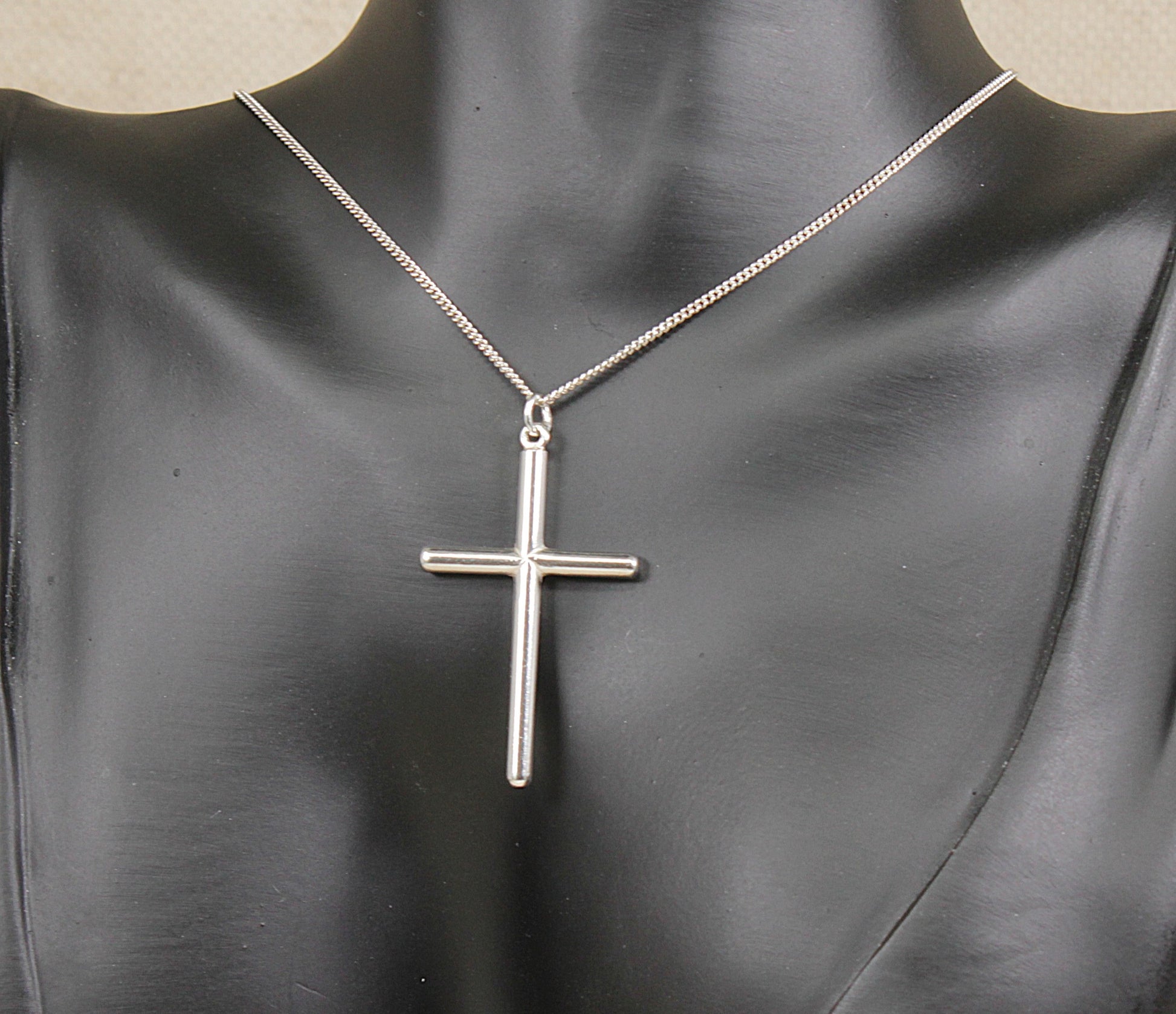 A silver cross pendant necklace displayed on a black mannequin bust against a light neutral background.