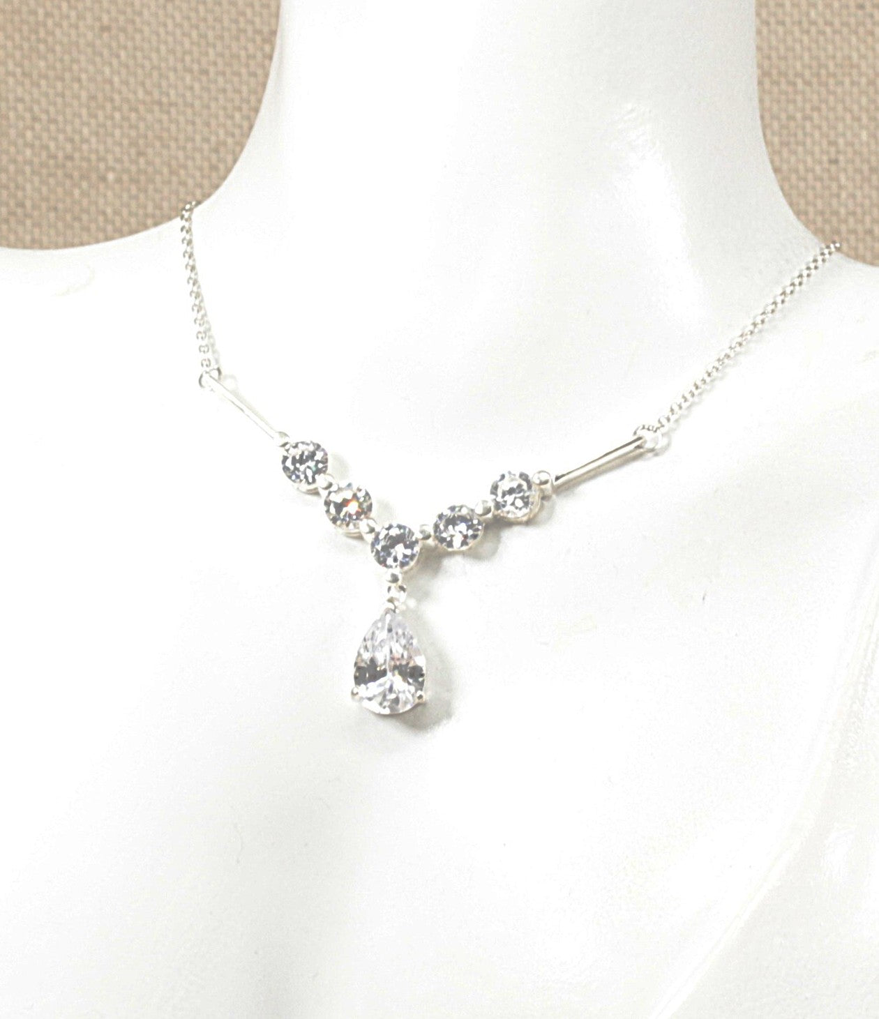 A silver necklace with a teardrop-shaped clear crystal pendant and smaller clear crystal beads along the chain, displayed on a white mannequin bust.