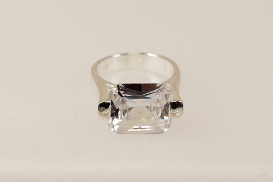 This silver CZ ring is a bold and striking piece, characterised by its substantial design and unique architectural setting.
The main feature is the large emerald cut Cubic Zirconia gemstone set in a cradle style mount, the stone is secured with 2 chunky claws that extend from the shoulders of the ring. The combination of the large CZ, heavy silver band, and innovative design makes this ring perfect for those who appreciate modern eye-catching statement jewellery. 

It is in excellent condition and inside t