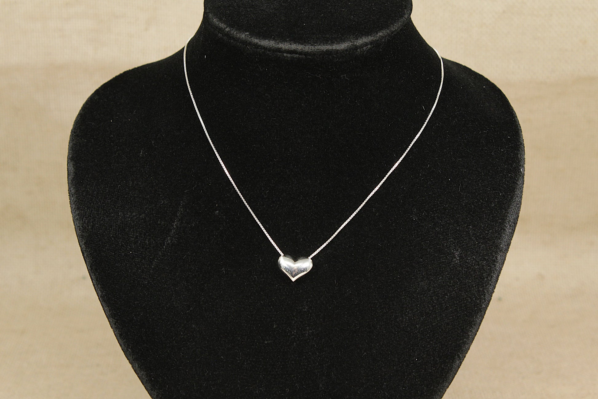 Silver heart-shaped pendant on a black velvet mannequin against a beige background