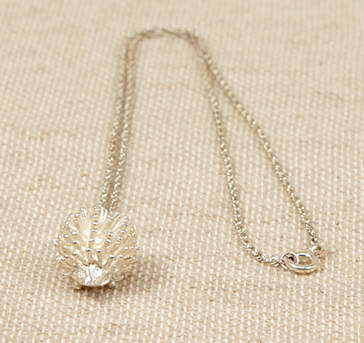 A sterling silver hedgehog pendant with a 'Prince of Wales' silver chain necklace laid out on a textured surface.