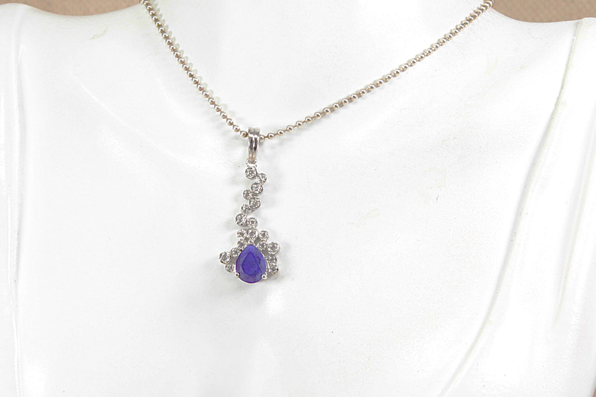 A silver chain necklace with a decorative pendant featuring a blue  teardrop gemstone surrounded by small clear stones, displayed on a white mannequin bust