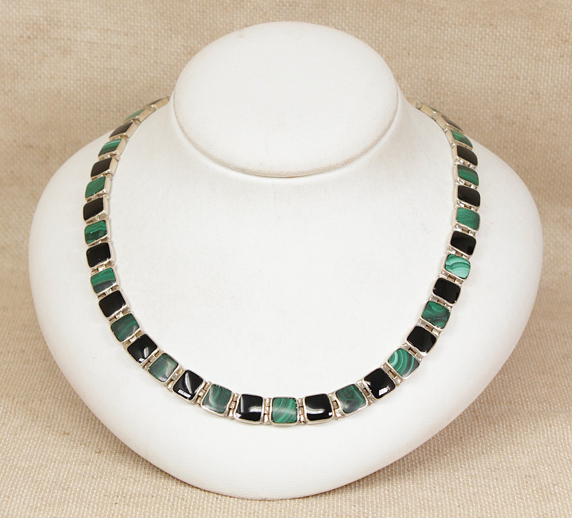 Necklace with green malachite and black onyx square gemstones on a white stand against a beige background