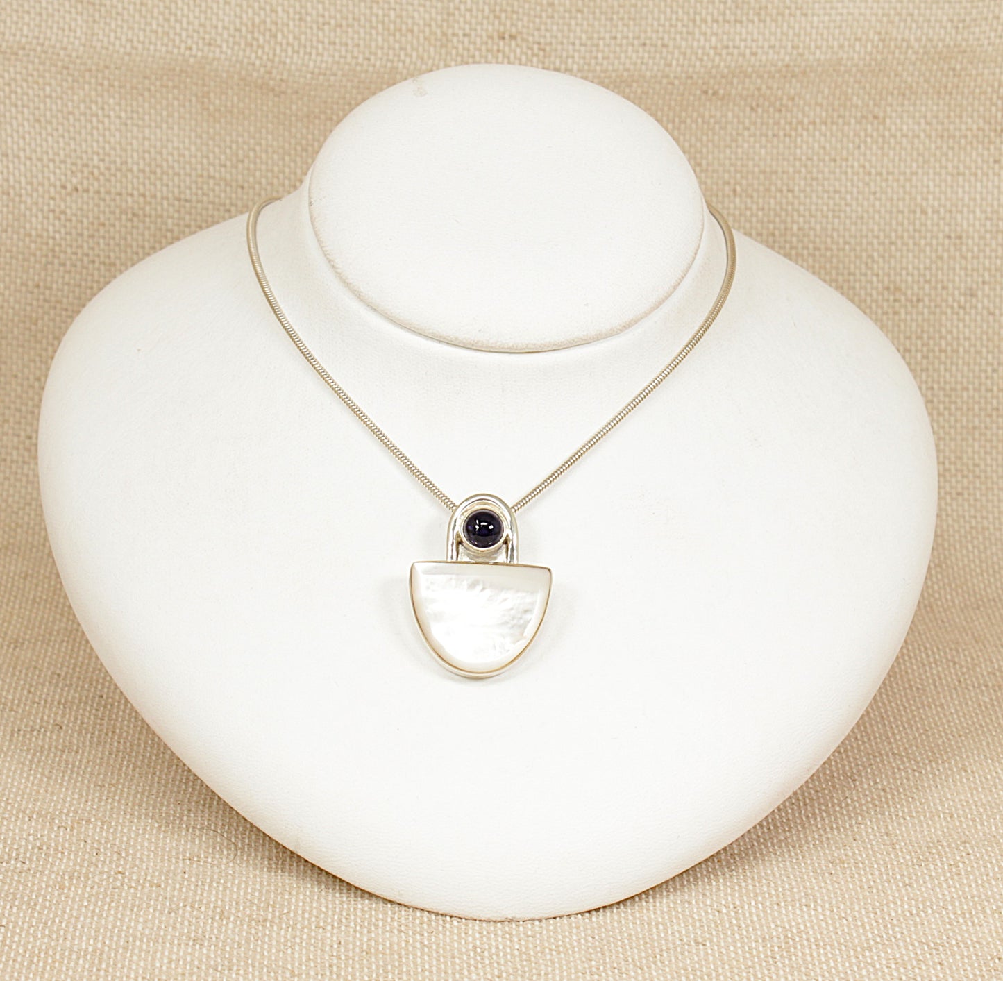 A striking and substantial sterling silver pendant showcasing a sleek, modernist design. The piece features a luminous semicircle of Mother of Pearl, bezel-set to highlight its natural iridescence. Above it rests a round cabochon of deep violet Iolite, adding a touch of rich colour and contrast to the composition. 
16 inches long.
