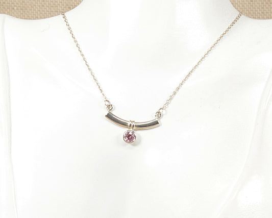 A silver necklace displayed on a white mannequin bust, featuring a curved bar pendant with a small round pink gemstone hanging from the center.