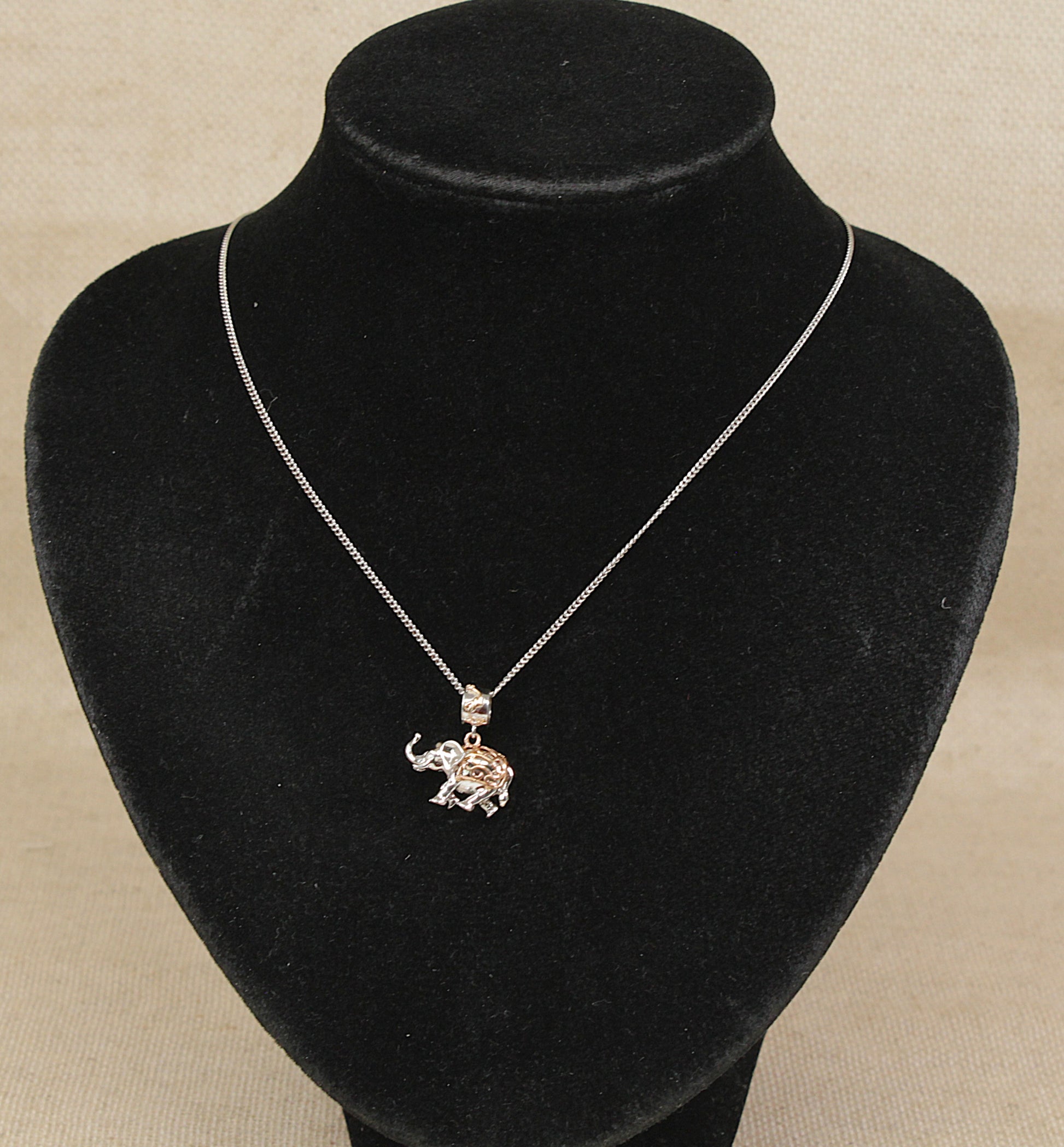 A silver chain necklace with a small silver and rare welsh gold elephant pendant, displayed on a black velvet jewelry bust against a light beige background.