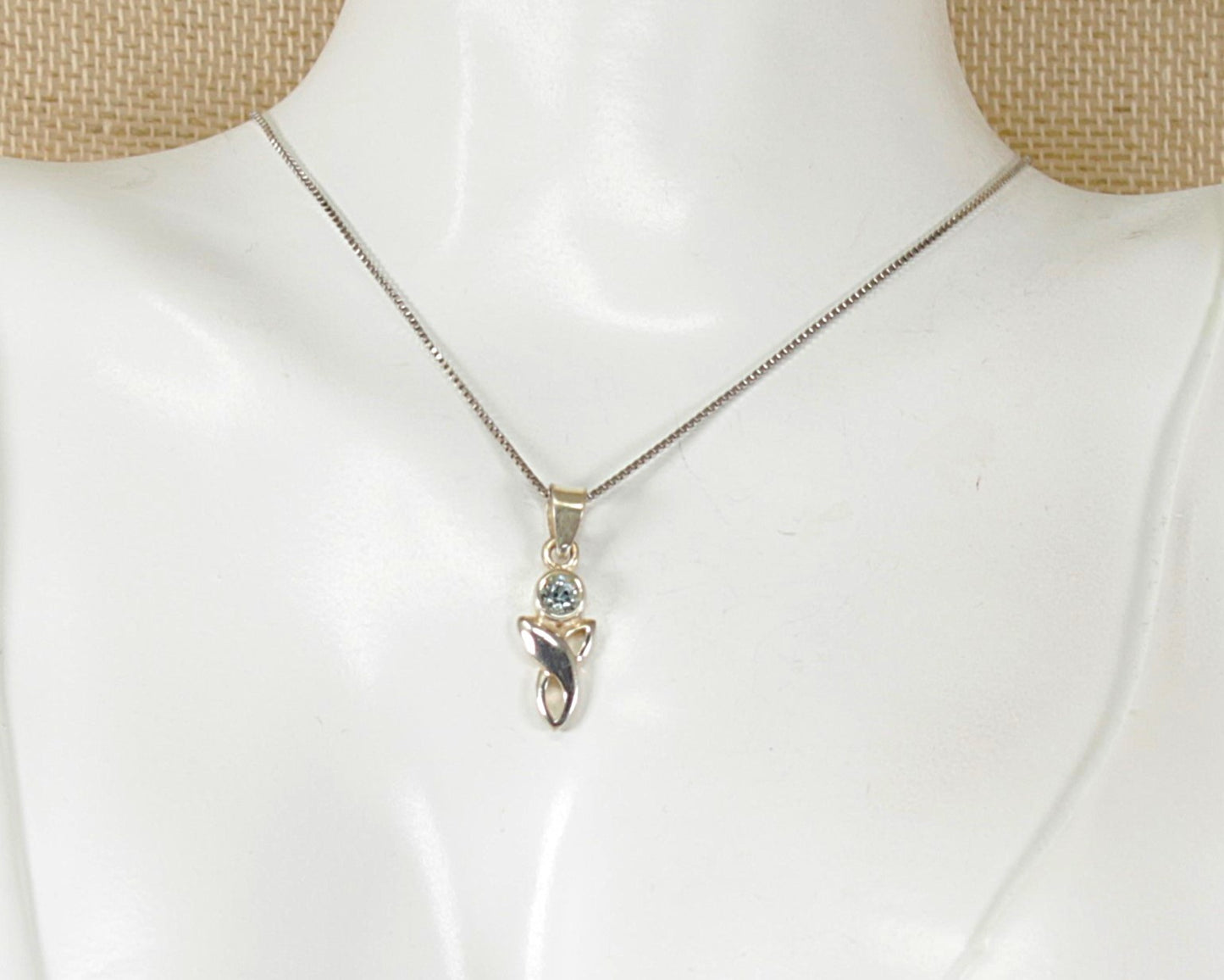 A silver chain necklace with a small pendant featuring a blue topaz gemstone and a silver celtic knot, displayed on a white mannequin bust.