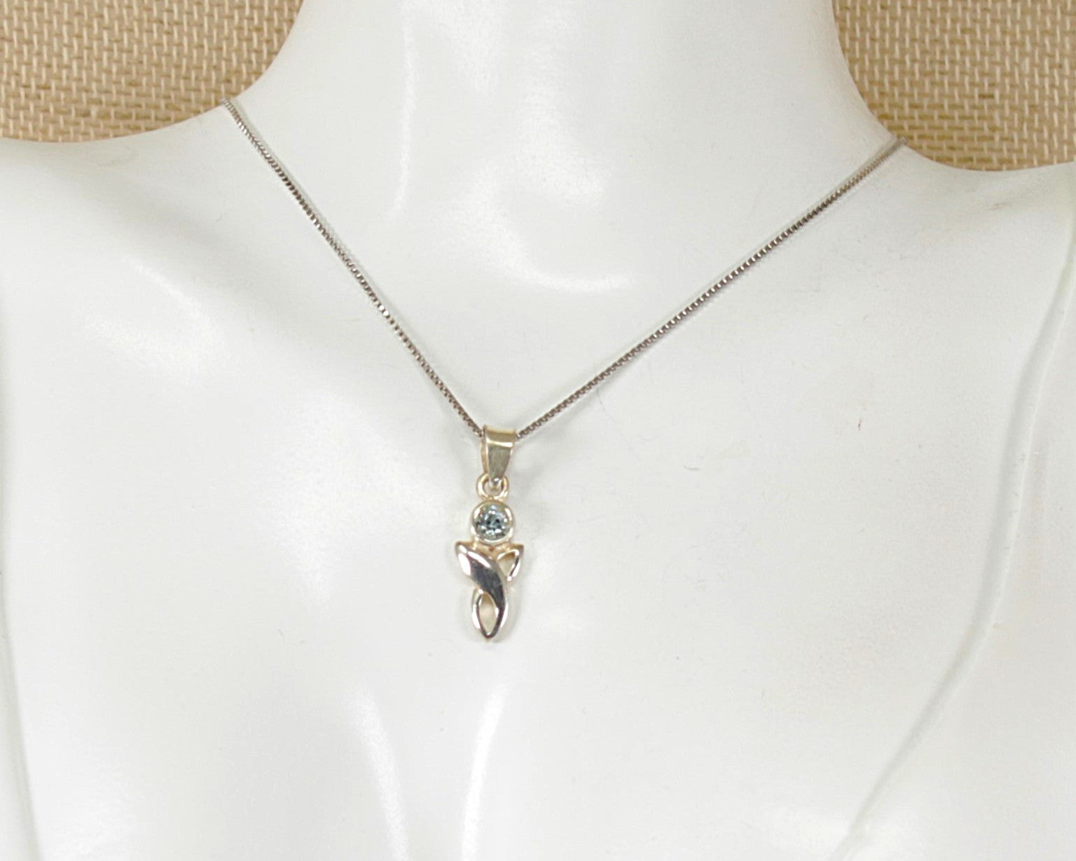 A silver chain necklace with a small pendant featuring a blue topaz gemstone and a silver celtic knot, displayed on a white mannequin bust.