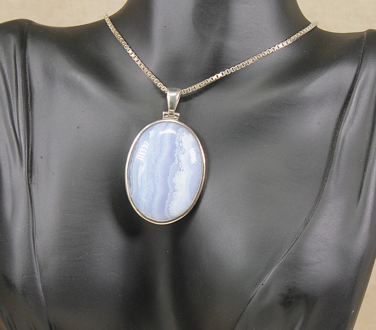An oval silver pendant necklace with a light blue lace agate stone, displayed on a black mannequin bust.