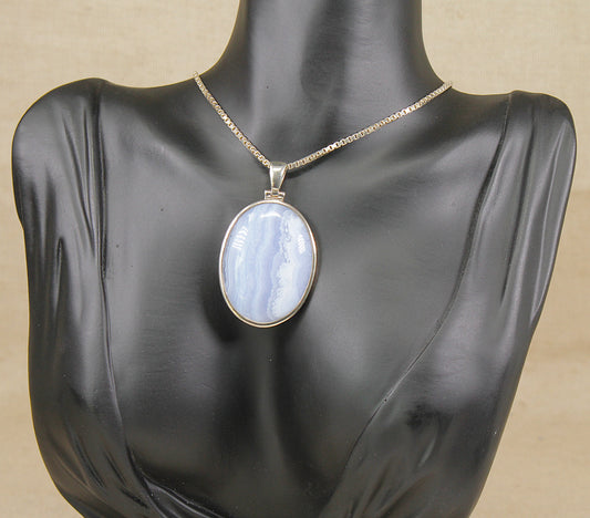 An oval pendant necklace with a light blue and white marbled agate stone in a silver bezel, displayed on a black mannequin bust against a beige background.