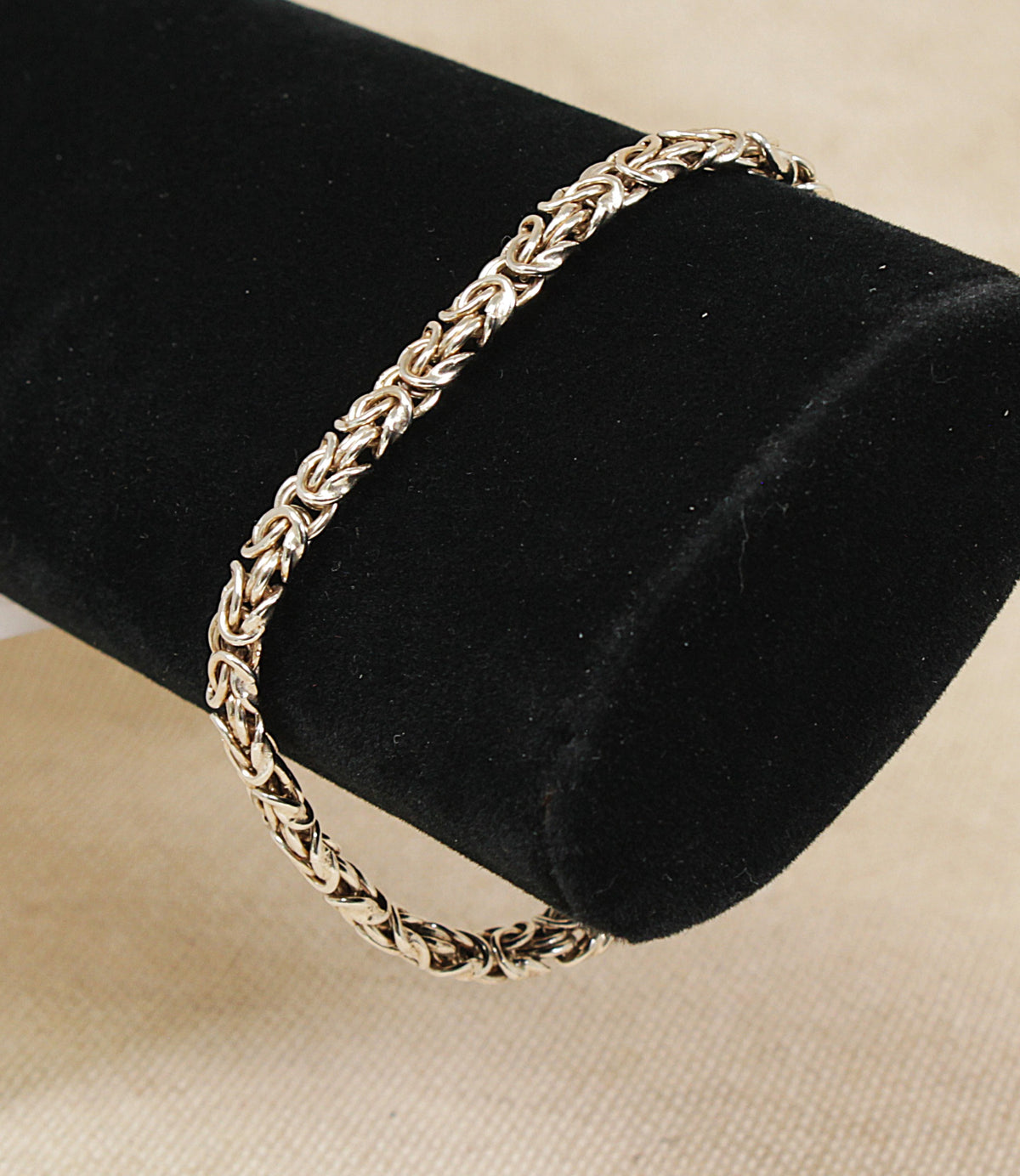 A silver-toned braided chain byzantine inspired bracelet displayed on a black velvet jewelry bust against a light beige fabric background.