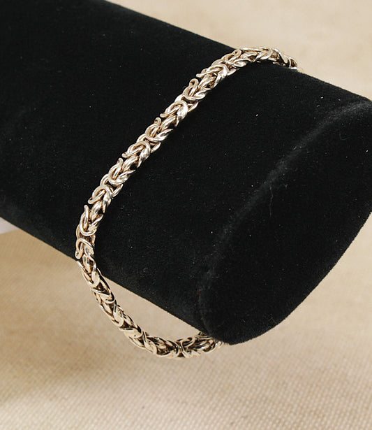 A silver-toned braided chain byzantine inspired bracelet displayed on a black velvet jewelry bust against a light beige fabric background.