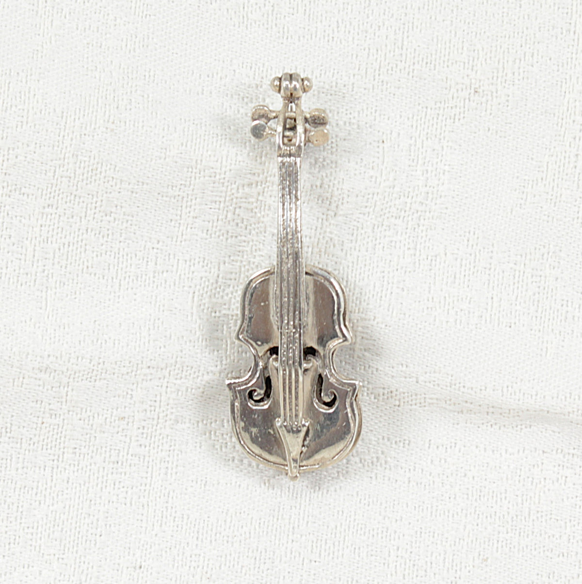 A silver cello-shaped brooch with an engraved scrollwork design on a textured white fabric background.