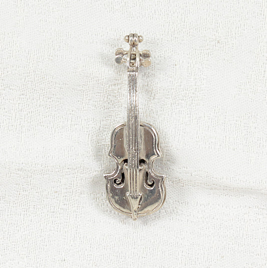 A silver cello-shaped brooch with an engraved scrollwork design on a textured white fabric background.