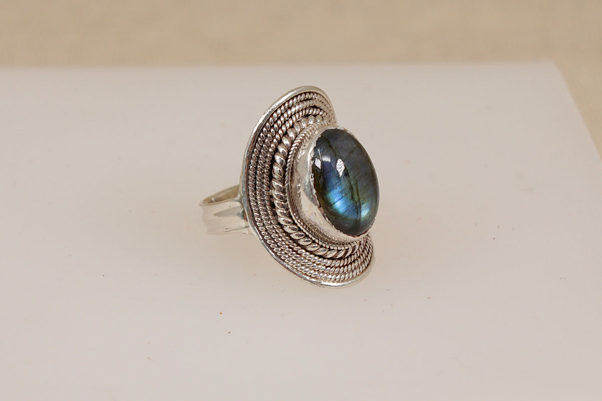 A suarti style silver ring featuring a large oval labradorite gemstone with a blue-green iridescent sheen, set in a textured, braided silver bezel.