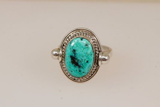A sterling silver ring featuring an oval turquoise stone with prominent black matrix, set in a decorative bezel with a twisted border and small side beads, shown on a plain light beige background.