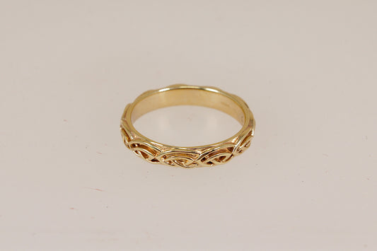 A yellow gold Annwyl band ring by Clogau with an intricate celtic pattern, shown on a plain light beige background.