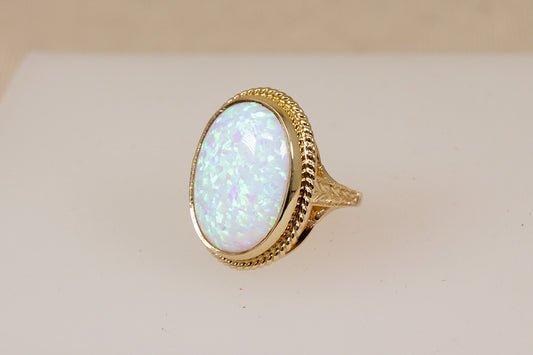A gold ring featuring an oval opal gemstone with iridescent blue and green flashes, set in a textured gold bezel on a light beige background.