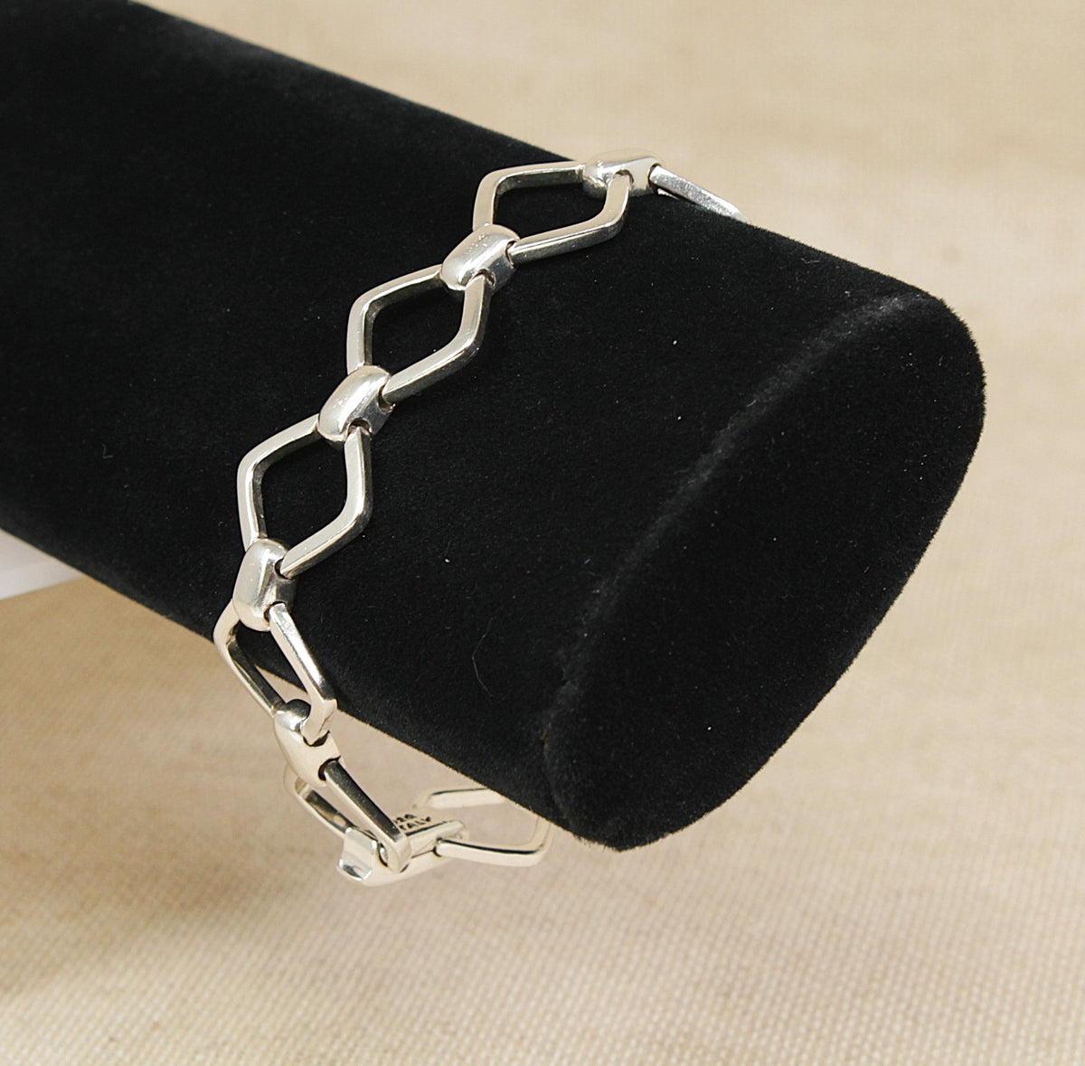A silver geometric link bracelet displayed on a black velvet cylindrical jewelry stand against a beige fabric background.