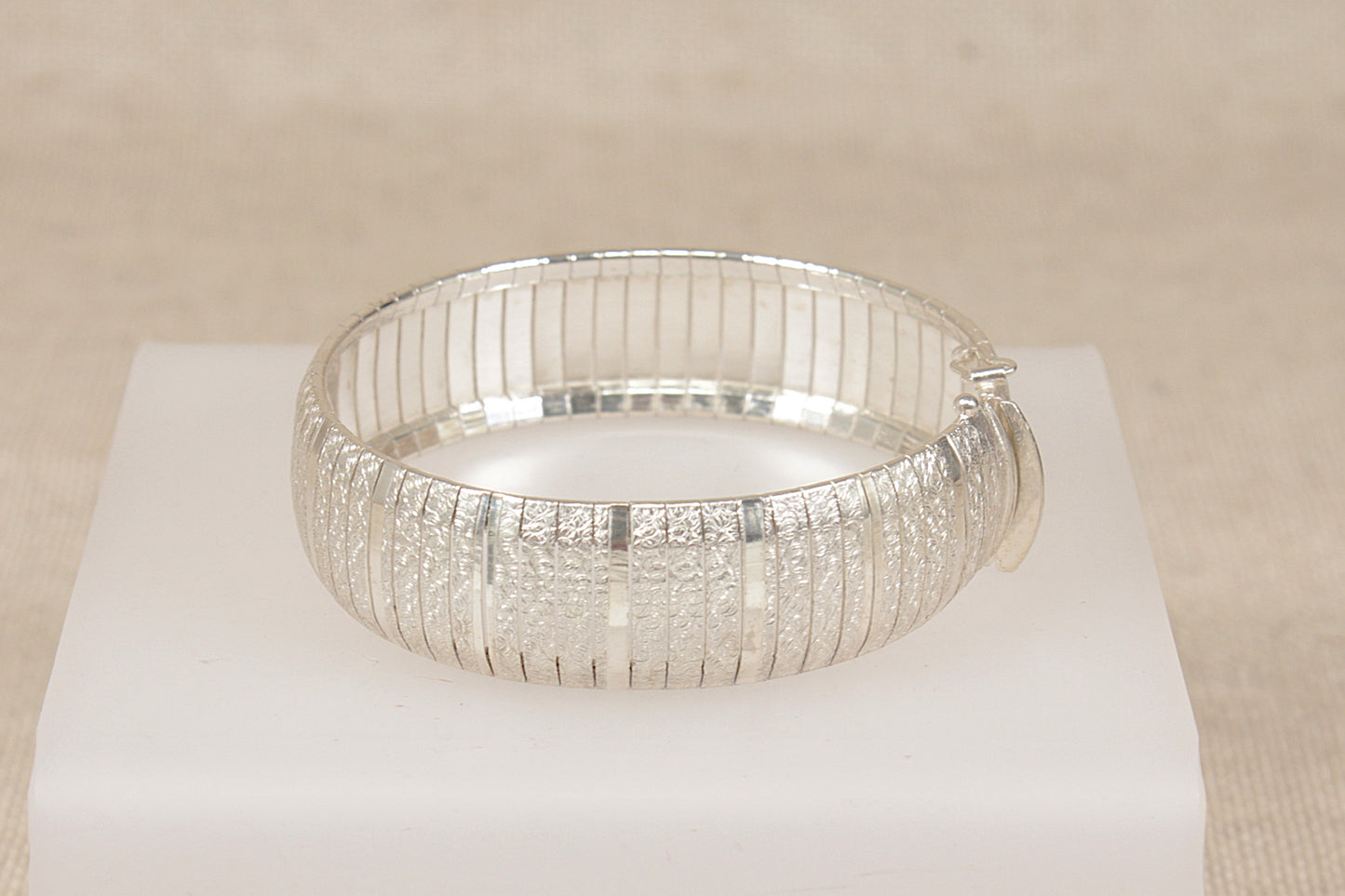 A silver textured bangle bracelet displayed on a white rectangular pedestal against a neutral beige background.